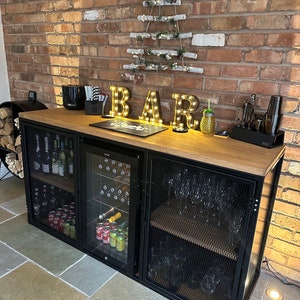 Puede incluir: Un bar de metal negro con una tapa de madera y puertas de vidrio. El bar tiene estantes para guardar botellas y vasos. El bar est&aacute; decorado con una cadena de luces que deletrea "BAR".
