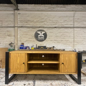 Puede incluir: Mueble de TV de madera clara con patas de metal negro. Tiene dos puertas, dos estantes y un logotipo circular "Polaris Handcrafted". El mueble est&aacute; en un taller, con una pared de ladrillo al fondo.