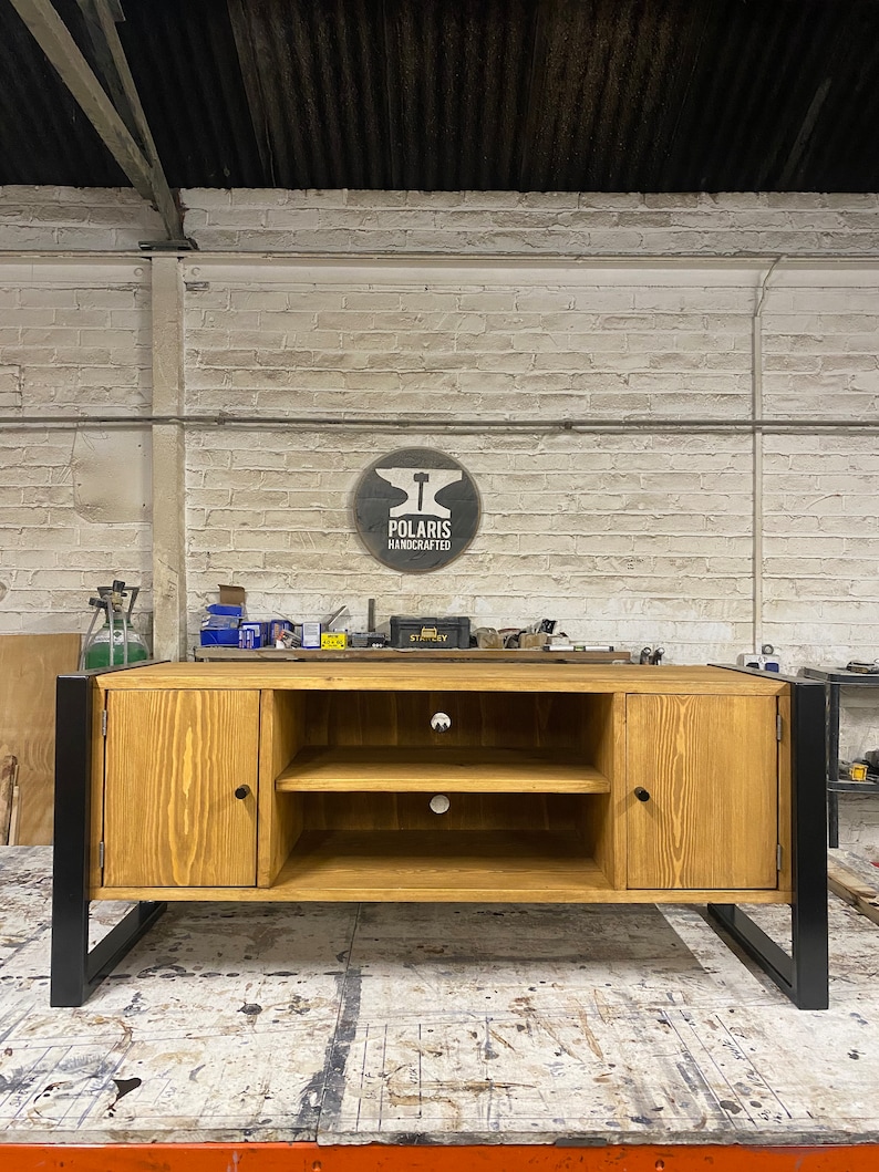 Puede incluir: Mueble de TV de madera clara con patas de metal negro. Tiene dos puertas, dos estantes y un logotipo circular "Polaris Handcrafted". El mueble est&aacute; en un taller, con una pared de ladrillo al fondo.