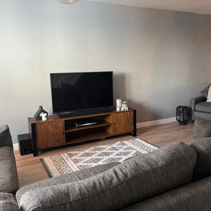 Puede incluir: Un sal&oacute;n moderno con un televisor de pantalla plana grande montado sobre una consola de medios de madera. Un sof&aacute; y un sill&oacute;n grises est&aacute;n dispuestos alrededor de una alfombra estampada. Los elementos decorativos incluyen velas, un jarr&oacute;n negro y una peque&ntilde;a planta. La habitaci&oacute;n tiene pisos de madera y paredes grises.