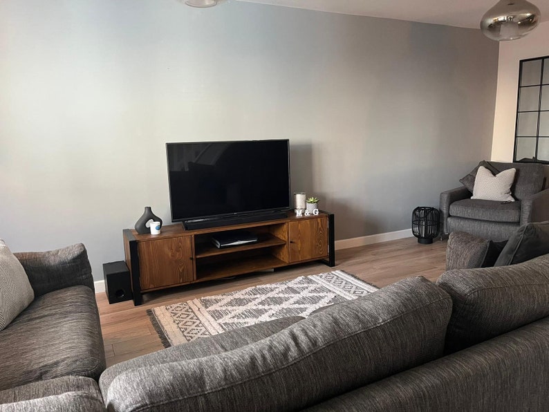 Puede incluir: Un sal&oacute;n moderno con un televisor de pantalla plana grande montado sobre una consola de medios de madera. Un sof&aacute; y un sill&oacute;n grises est&aacute;n dispuestos alrededor de una alfombra estampada. Los elementos decorativos incluyen velas, un jarr&oacute;n negro y una peque&ntilde;a planta. La habitaci&oacute;n tiene pisos de madera y paredes grises.