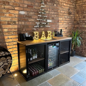 Puede incluir: Un armario de bar de metal negro con encimera de madera y puertas de malla met&aacute;lica. El armario tiene un refrigerador incorporado y est&aacute; lleno de botellas de vino y latas. El armario est&aacute; frente a una pared de ladrillo con un &aacute;rbol de Navidad decorativo iluminado.