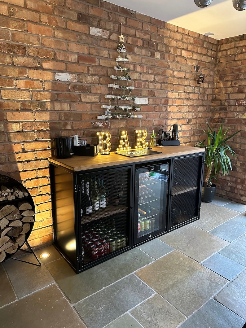 Puede incluir: Un armario de bar de metal negro con encimera de madera y puertas de malla met&aacute;lica. El armario tiene un refrigerador incorporado y est&aacute; lleno de botellas de vino y latas. El armario est&aacute; frente a una pared de ladrillo con un &aacute;rbol de Navidad decorativo iluminado.