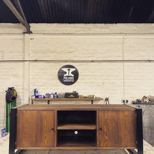 Puede incluir: Un mueble de TV de madera artesanal con acabado natural y patas de metal negro. El mueble tiene dos puertas y un estante abierto. El letrero de arriba dice "POLARIS HANDCRAFTED".