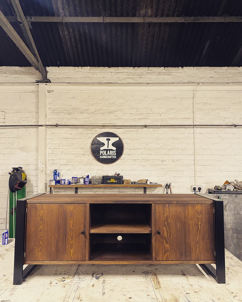 Puede incluir: Un mueble de TV de madera artesanal con acabado natural y patas de metal negro. El mueble tiene dos puertas y un estante abierto. El letrero de arriba dice "POLARIS HANDCRAFTED".