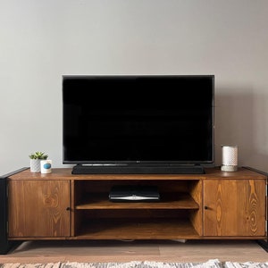Puede incluir: Un mueble de TV de madera con marco de metal negro, que sostiene un televisor de pantalla plana grande. El mueble tiene dos puertas y estantes abiertos, con una peque&ntilde;a planta y una vela encima. Tambi&eacute;n se ve una barra de sonido negra y un subwoofer.