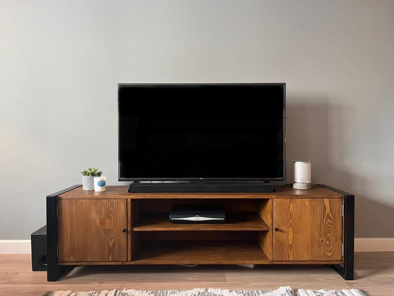 Puede incluir: Un mueble de TV de madera con marco de metal negro, que sostiene un televisor de pantalla plana grande. El mueble tiene dos puertas y estantes abiertos, con una peque&ntilde;a planta y una vela encima. Tambi&eacute;n se ve una barra de sonido negra y un subwoofer.