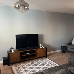 Puede incluir: Un sal&oacute;n moderno con un televisor de pantalla plana grande sobre un mueble de televisi&oacute;n de madera. La habitaci&oacute;n cuenta con un sof&aacute; gris, un sill&oacute;n y una alfombra estampada. Dos l&aacute;mparas colgantes plateadas cuelgan del techo y las paredes est&aacute;n pintadas de gris claro.