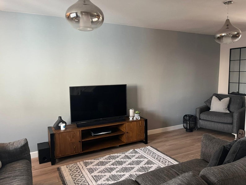 Puede incluir: Un sal&oacute;n moderno con un televisor de pantalla plana grande sobre un mueble de televisi&oacute;n de madera. La habitaci&oacute;n cuenta con un sof&aacute; gris, un sill&oacute;n y una alfombra estampada. Dos l&aacute;mparas colgantes plateadas cuelgan del techo y las paredes est&aacute;n pintadas de gris claro.