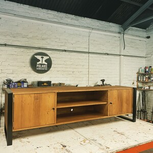 Puede incluir: Mueble de TV de madera artesanal con acabado natural y patas de metal negro. Cuenta con dos puertas, estanter&iacute;as abiertas y un orificio para la gesti&oacute;n de cables. El logotipo de Polaris Handcrafted es visible. Mide unos 180 cm de largo.