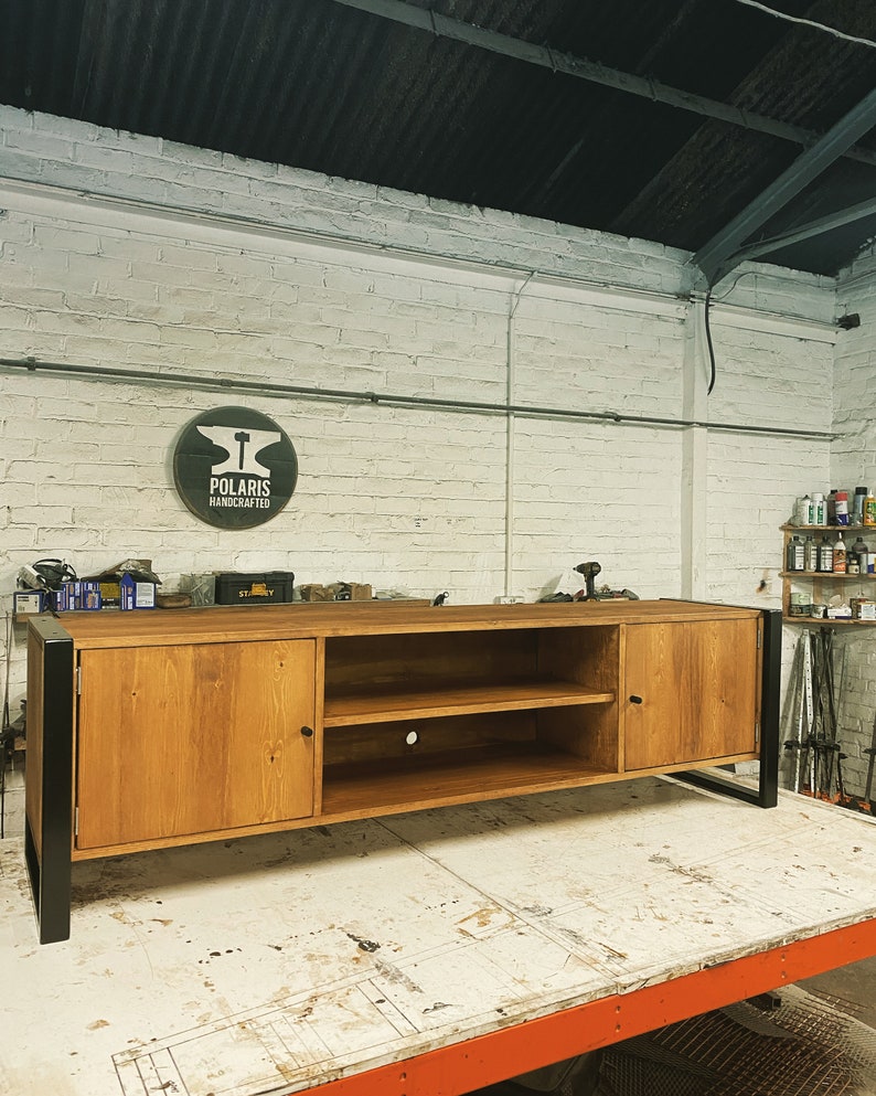 Puede incluir: Mueble de TV de madera artesanal con acabado natural y patas de metal negro. Cuenta con dos puertas, estanter&iacute;as abiertas y un orificio para la gesti&oacute;n de cables. El logotipo de Polaris Handcrafted es visible. Mide unos 180 cm de largo.