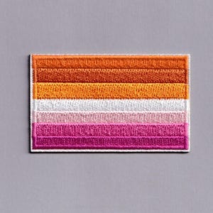 May include: A rectangular embroidered patch featuring five horizontal stripes in shades of orange, light orange, white, light pink, and dark pink. The patch has a visible stitched border and a textured fabric surface with a consistent thread pattern.