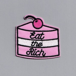 Könnte beinhalten: Rosa gestickter Bügel-Patch in Form eines Kuchens mit einer Kirsche oben drauf. Der Kuchen hat weißen Zuckerguss und den Text "Eat the Rich" in Schwarz.