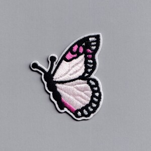 Puede incluir: Un parche bordado con una mariposa blanca con detalles rosas y contornos negros. La mariposa está mirando hacia la derecha y tiene las alas extendidas.
