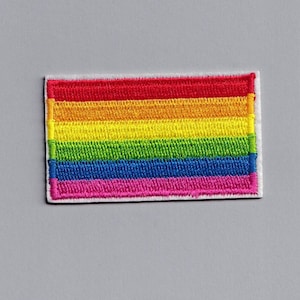 Puede incluir: Un parche de bandera del orgullo arcoíris con seis rayas horizontales en los colores rojo, naranja, amarillo, verde, azul y rosa. El parche está bordado sobre un fondo de tela blanco.