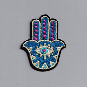 May include: An embroidered hamsa hand in blue and white with a pink eye in the centre. The hamsa is a symbol of protection and good luck.