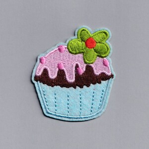 Può includere: Un cupcake blu con glassa rosa e un fiore verde in cima. Il cupcake ha una glassa al cioccolato marrone.