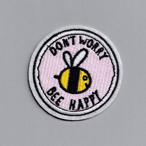 May include: A round embroidered patch with a pink background and white border. The patch features a cartoon bee with black and yellow stripes and the text "Don't Worry Bee Happy".
