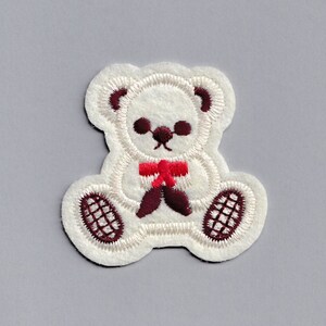 Peut inclure: Applique en feutre blanc d'un ours en peluche avec un nœud rouge. L'ours est assis, les pattes devant lui. Les yeux, le nez et la bouche de l'ours sont brodés en fil brun. Les pattes de l'ours sont brodées en fil brun avec un motif en croisillons.