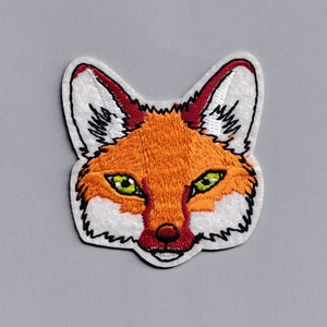 Peut inclure: Patch brodé représentant la tête d'un renard avec une fourrure orange, un museau blanc et des yeux verts.