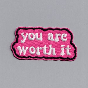 Può includere: Toppa ricamata rosa con le parole "you are worth it" in bianco, bordate di nero. La toppa ha una forma a nuvola ed è posta su uno sfondo grigio.