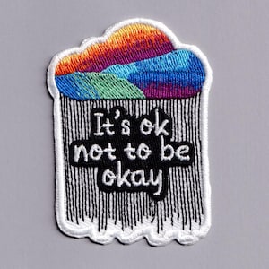 May include: An embroidered patch featuring a colourful cloud with orange, purple, and blue gradients at the top. Below the cloud, black vertical lines represent rain, framing the white embroidered text that reads, It's ok not to be okay.