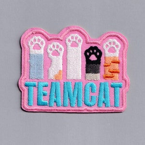May include: Pink embroidered patch with five paw prints in different colors and the text "TEAMCAT" in blue.