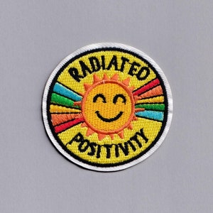 Puede incluir: Un parche bordado amarillo con un sol sonriente y el texto "RADIATED POSITIVITY" en negro.