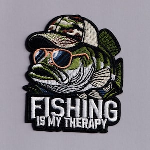 Puede incluir: Un parche bordado que muestra un pez lubina verde con una gorra de béisbol de camuflaje y gafas de sol de montura marrón. Debajo del pez, el texto FISHING IS MY THERAPY aparece en letras mayúsculas blancas sobre un borde de fondo negro.