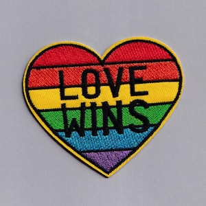 Op de afbeelding: Een hartvormige patch met een regenboogontwerp en de woorden "LOVE WINS" geborduurd in zwart. De patch heeft een gele rand en is geplaatst tegen een grijze achtergrond. De kleuren van de regenboog zijn rood, oranje, geel, groen, blauw en paars.