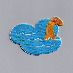 May include: An embroidered patch of a brown sea monster with a pink nose, swimming in blue water with green fish. The patch is shaped like a cloud.