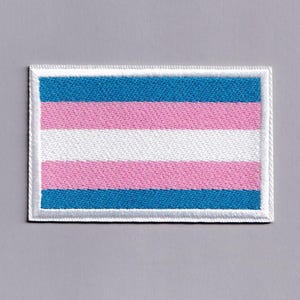 Op de afbeelding: Een rechthoekige geborduurde patch met het ontwerp van de transgender pride-vlag. De patch heeft horizontale strepen in lichtblauw, roze, wit, roze en lichtblauw, allemaal omsloten door een dikke, effen witte geborduurde rand.