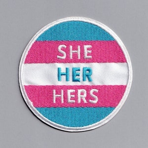 Op de afbeelding: Een ronde, geborduurde patch met de kleuren van de transgender pride-vlag. De patch heeft de tekst "SHE HER HERS" in wit.