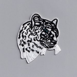 Pode incluir: Um remendo bordado da cabeça de um leopardo das neves, com pelagem branca e preta e um olho azul.
