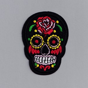 May include: Black embroidered iron-on patch of a sugar skull with a red rose, yellow and green floral details, and white teeth.