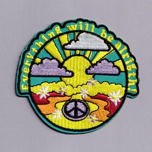 May include: A circular embroidered patch featuring a yellow sun, purple clouds, and a white cloud against a teal sky. The foreground shows orange hills, white flowers, and a purple peace sign. The text Everything will be alright is embroidered in yellow.