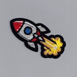 May include: An embroidered patch featuring a cartoon rocket ship in black and white with red and blue accents. The rocket is launching with a yellow flame.