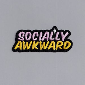 Op de afbeelding: Een zwarte geborduurde patch met de tekst "SOCIALLY AWKWARD" in roze en gele letters.