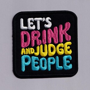 May include: A square embroidered patch with a black background and rounded corners. The text reads LET'S in white, DRINK in pink, AND in yellow, JUDGE in yellow, and PEOPLE in light blue. The embroidery features a raised, textured stitch.