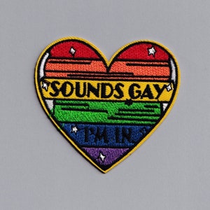 Puede incluir: Un parche en forma de corazón arcoíris con el texto "SOUNDS GAY I'M IN" en letras negras y amarillas.
