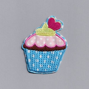 Op de afbeelding: Een blauwe en roze cupcake met witte stippen en een roze hart bovenop. De cupcake is geborduurd op een stoffen patch.