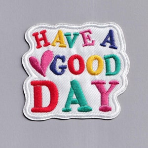 May include: White fabric patch with colorful embroidered text that reads "Have a good day" with a pink heart in the middle of the word "good".