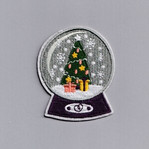 May include: Embroidered patch with a snow globe design. Inside the globe is a green Christmas tree decorated with pink and yellow ornaments, and gifts. The base of the snow globe is purple, and the globe is clear with white snowflakes.