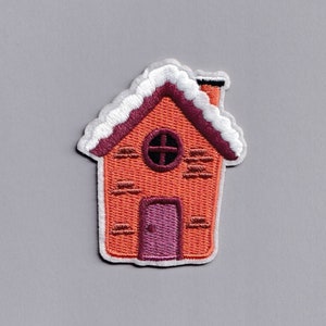 May include: Embroidered patch of a house with a red brick facade, a purple door, and a round window. The roof is burgundy and topped with white snow. A black chimney is visible.