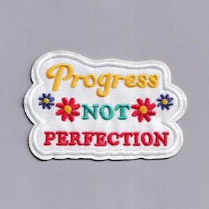 Pode incluir: Etiqueta branca bordada com o texto "Progress not perfection" em letras amarelas, verdes e vermelhas. A etiqueta é cercada por quatro flores coloridas.