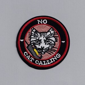 May include: A round, red and black embroidered patch with a white cat inside. The cat is holding a phone to its ear and the text "NO CAT CALLING" is around the cat.