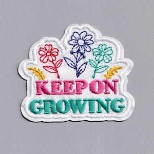 May include: White embroidered patch with colourful flowers and the text "KEEP ON GROWING" in pink and teal.