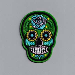 May include: Green embroidered skull with a blue and pink flower on the top, surrounded by yellow and blue details.