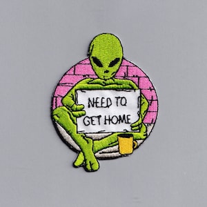 Könnte beinhalten: Gestickter Aufnäher mit einem grünen Alien, das ein Schild mit der Aufschrift "NEED TO GET HOME" hält. Das Alien sitzt vor einer rosa Backsteinmauer, mit einer gelben Tasse daneben. Der Aufnäher hat einen schwarzen Rand und befindet sich auf grauem Hintergrund.