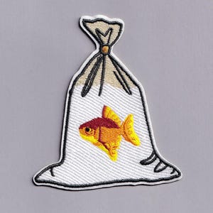 May include: An embroidered patch featuring a goldfish inside a clear plastic bag. The fish is orange and yellow with a dark red head. The bag is white with black outlines, tied at the top with a tan knot, and has a subtle wavy texture throughout.